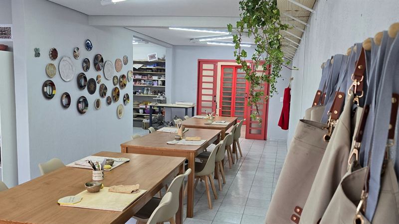 Mesas de trabalho do ateliê com pratos de cerâmica decorados na parede e prateleiras ao fundo