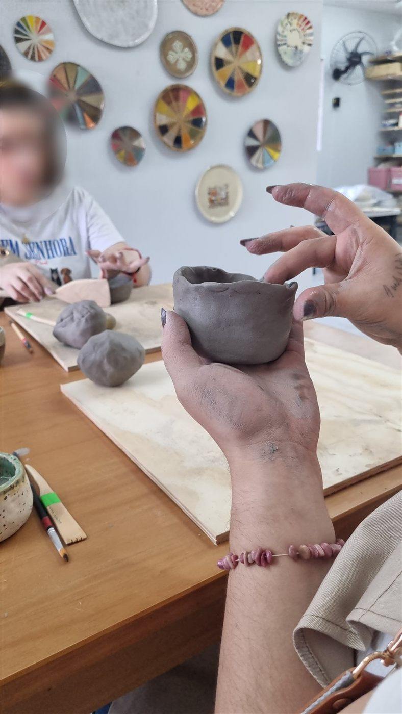 Mãos segurando uma cumbuca de argila, com outra participante trabalhando ao fundo