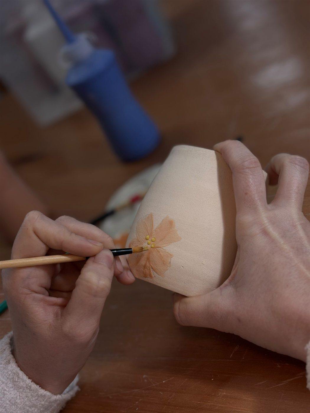 Mão pintando uma flor laranja em um vaso de cerâmica ainda sem esmalte